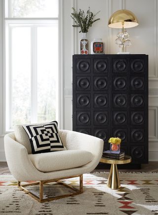 living room with cream boucle armchair, patterned rug, gold side table, black tall boy, gold table lamp, white walls