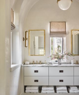 A cream bathroom with marble-topped double vanity unit and gold-framed mirrors