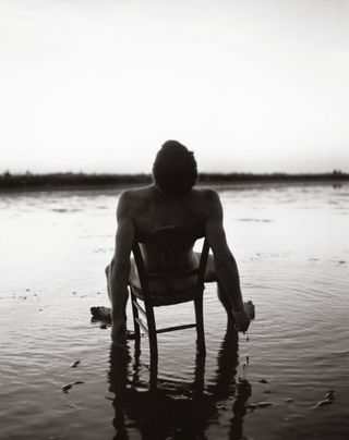 black and white photograph showing a nude man sitting on a chair in water