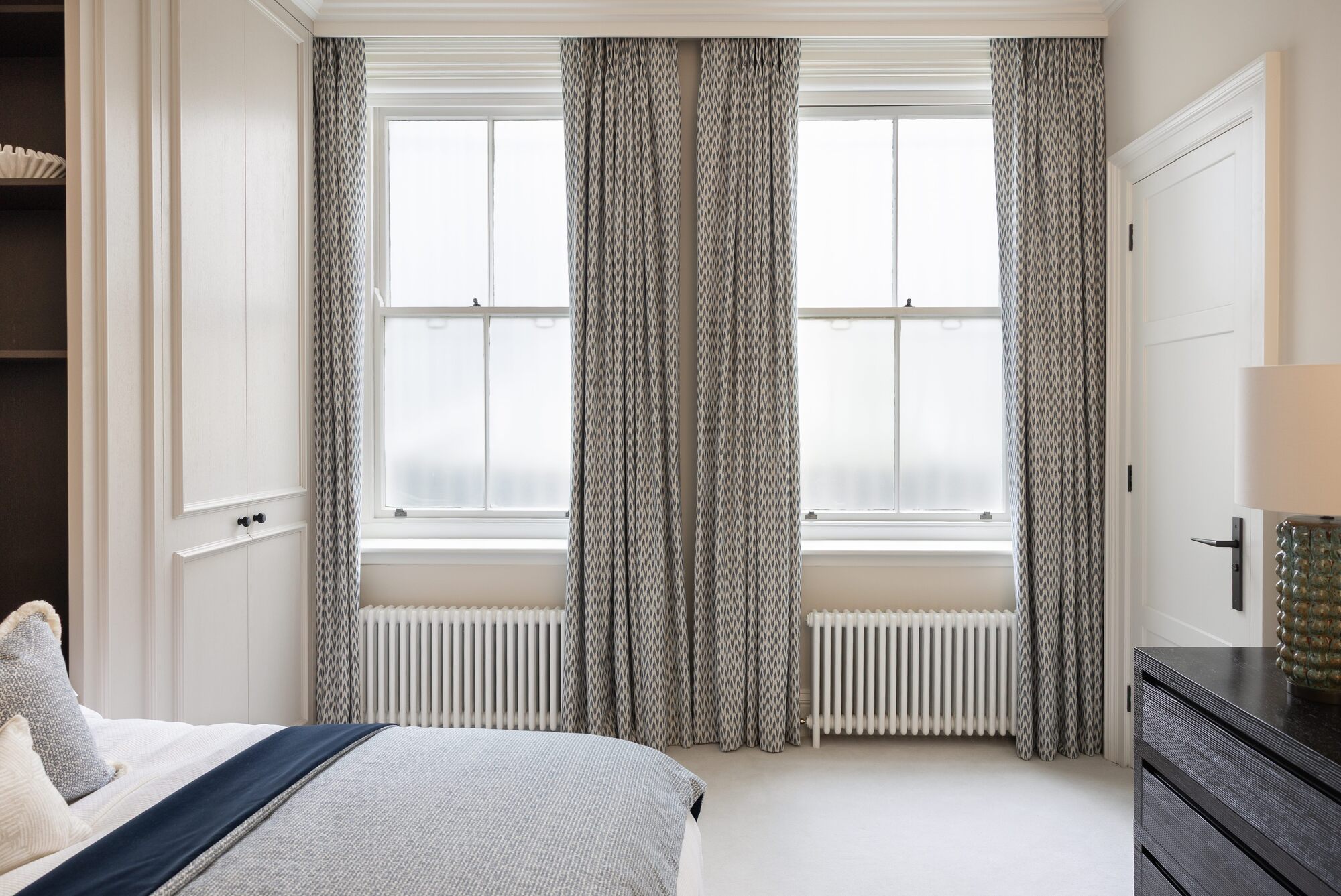 modern bedroom with two windows with curtains and radiators, built-in wardrobes with bed, black console with lamp on it