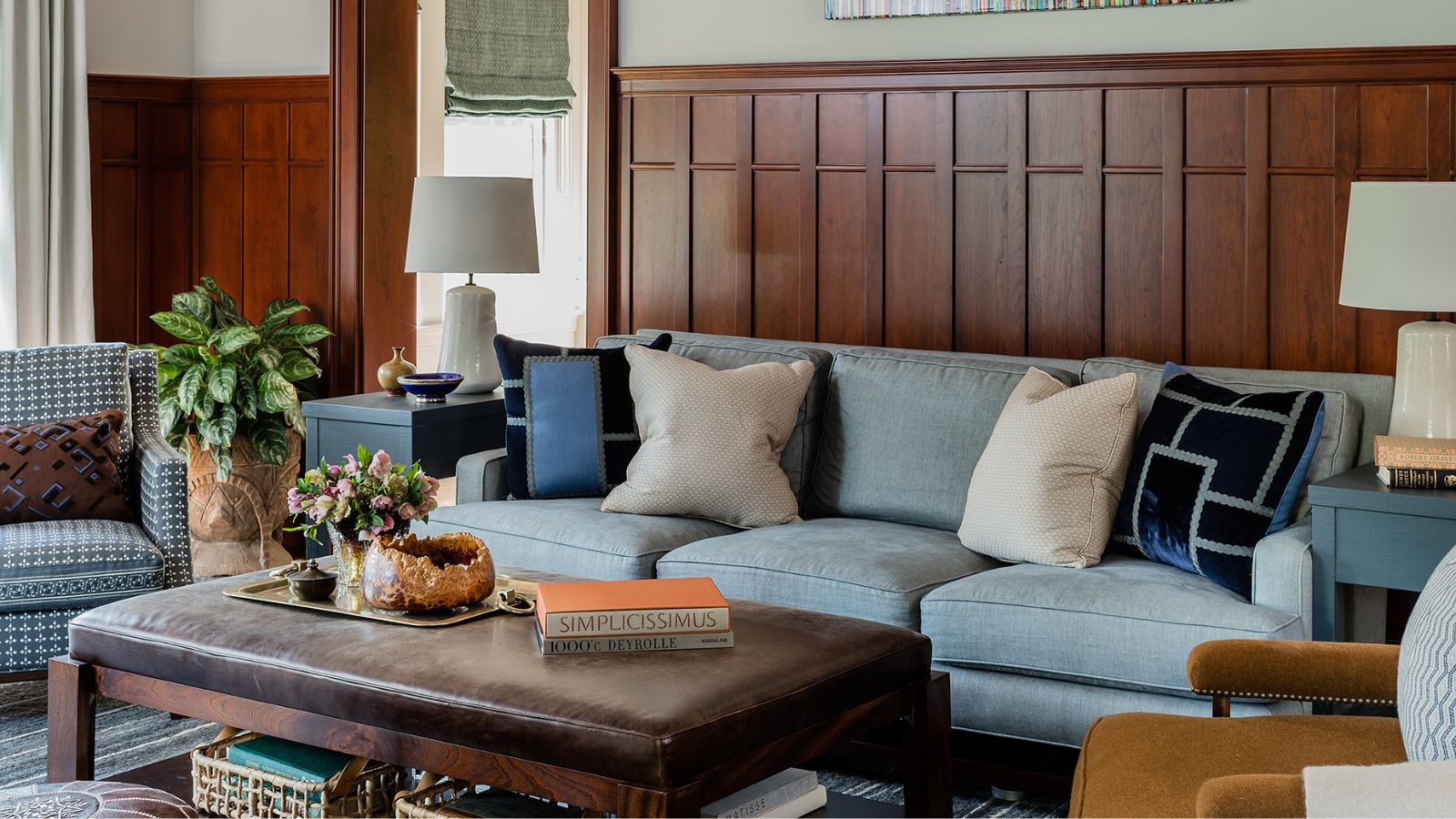 Living room with dark wood panelled walls, a pale blue sofa at the centre with cushions on top, a dark brown leather coffee table, two leather pouffes underneath, and a mustard velvet armchair.