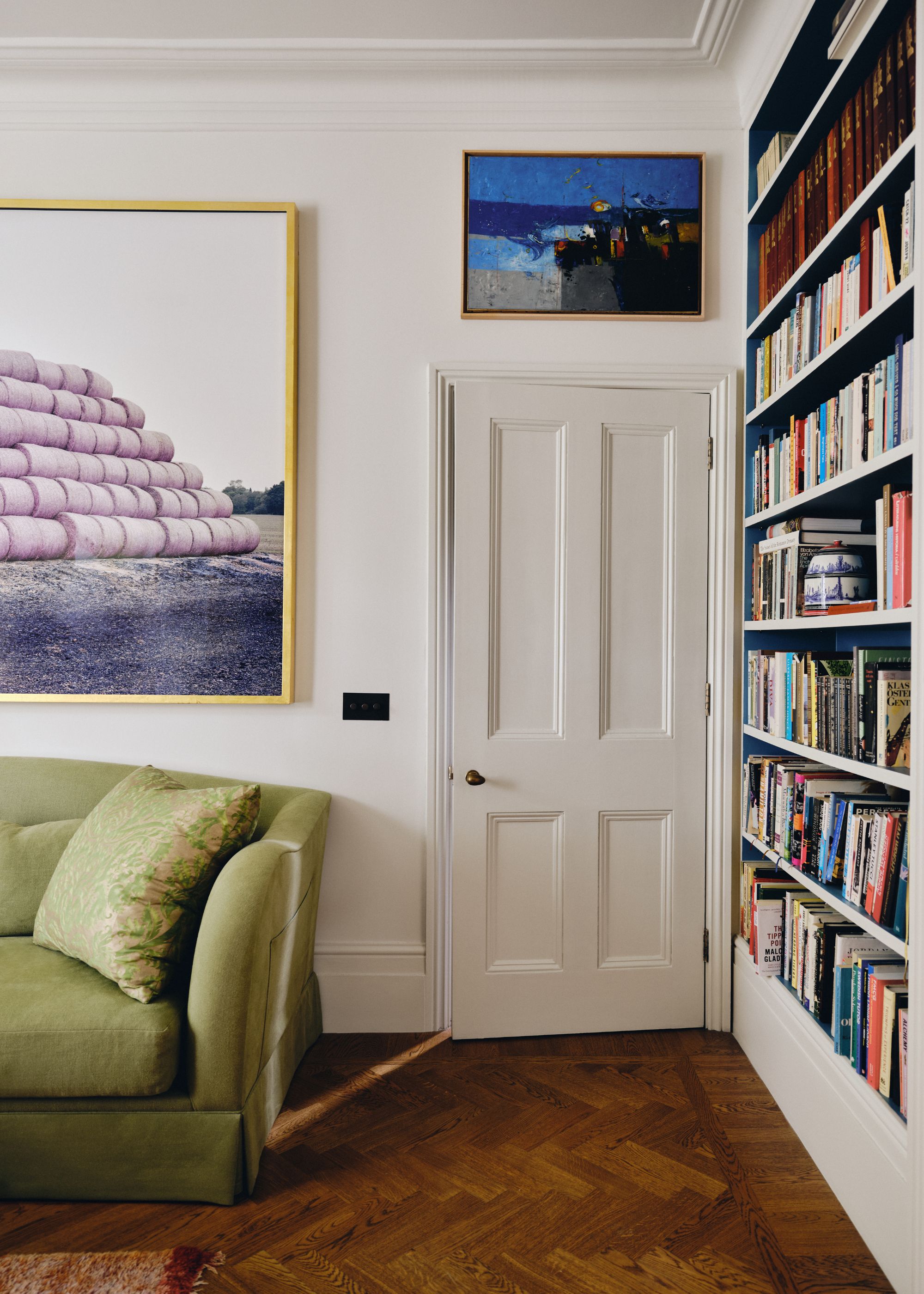 A living room with white walls, parquet flooring, a lime green sofa, and lilac artwork above it. Bookshelves on the other wall.
