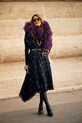 A woman at Paris Fashion Fall 2025 week wearing the purple color trend in a black and purple plaid dress, purple feathered boa, black shirt, and black boots