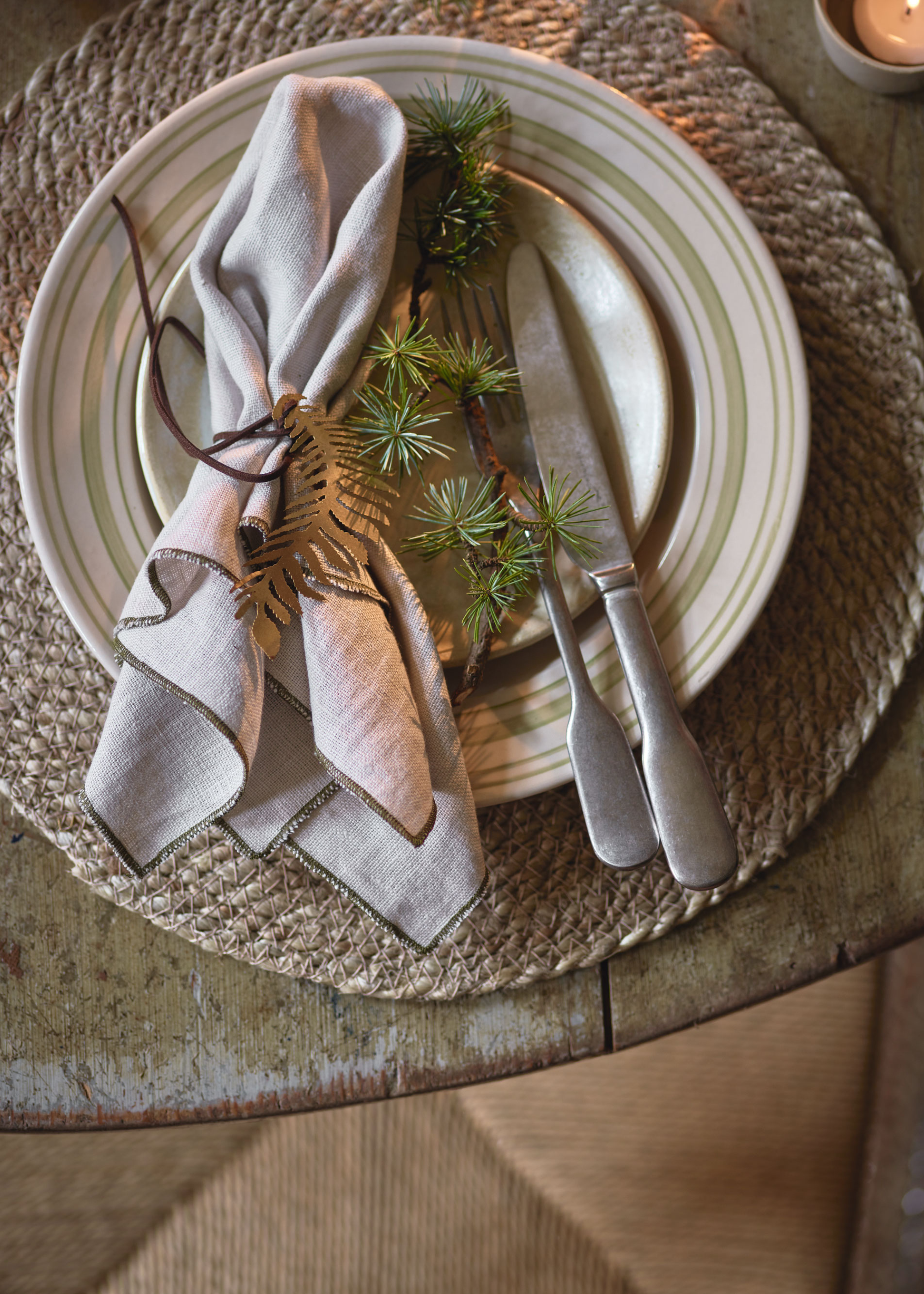 A Christmas table place setting with a knife and fork on a side plate that sits on top of a finner plate on a round placemat, with some foliage placed on the plate next to a napkin tied with string