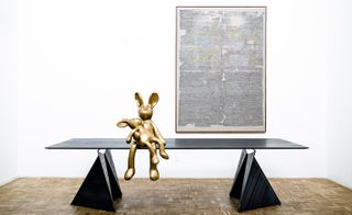 View of framed art on the wall, a black table with triangular legs and two bronze rabbit-like characters sitting on the edge of the table in a space with white walls and brown patterned flooring