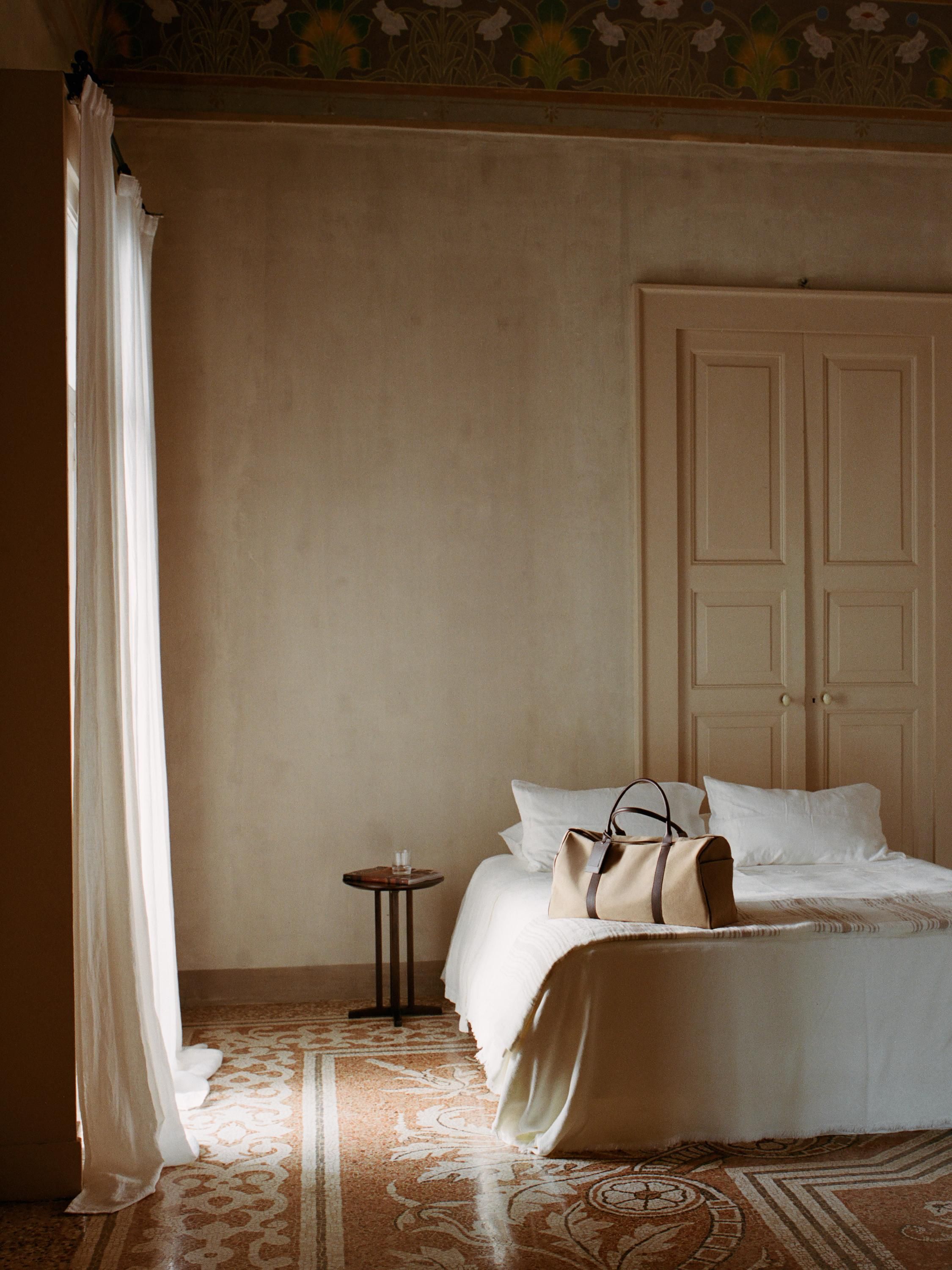 Image of a tan canvas travel suitcase on a white bed in a beige room with high ceilings and tiled flooring.