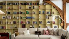 Living room with beamed ceiling and walls and a large built-in bookcase painted yellow, filled with books in each alcove. A large cream sectional sofa sits in the middle with patterned pillows.