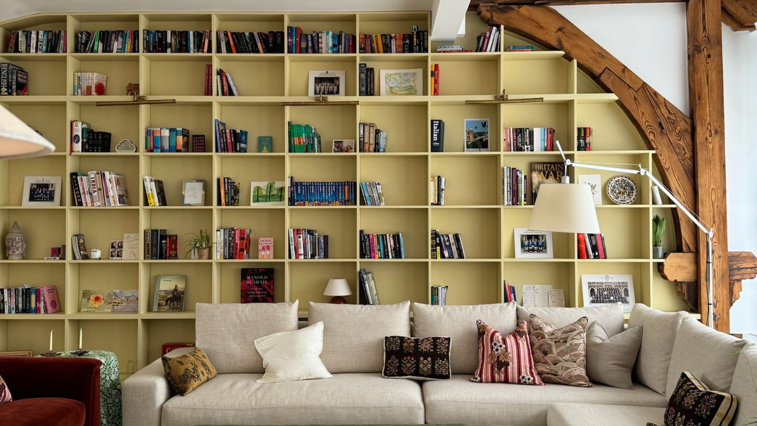 Living room with beamed ceiling and walls and a large built-in bookcase painted yellow, filled with books in each alcove. A large cream sectional sofa sits in the middle with patterned pillows.