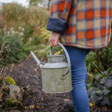 A woman in flannel walks through a garden holding a metal watering can