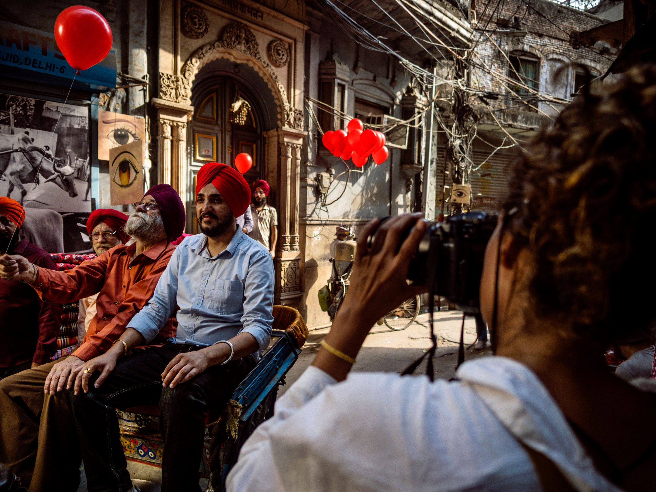 A female photographer placed to the right is photographing three men with red and black turbans sitting on chairs with red balloons in the background