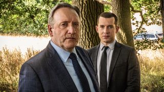 Neil Dudgeon and Nick Hendrik in suits as DCI John Barnaby and DS Jamie Winter stand in front of a tree in Midsomer Murders