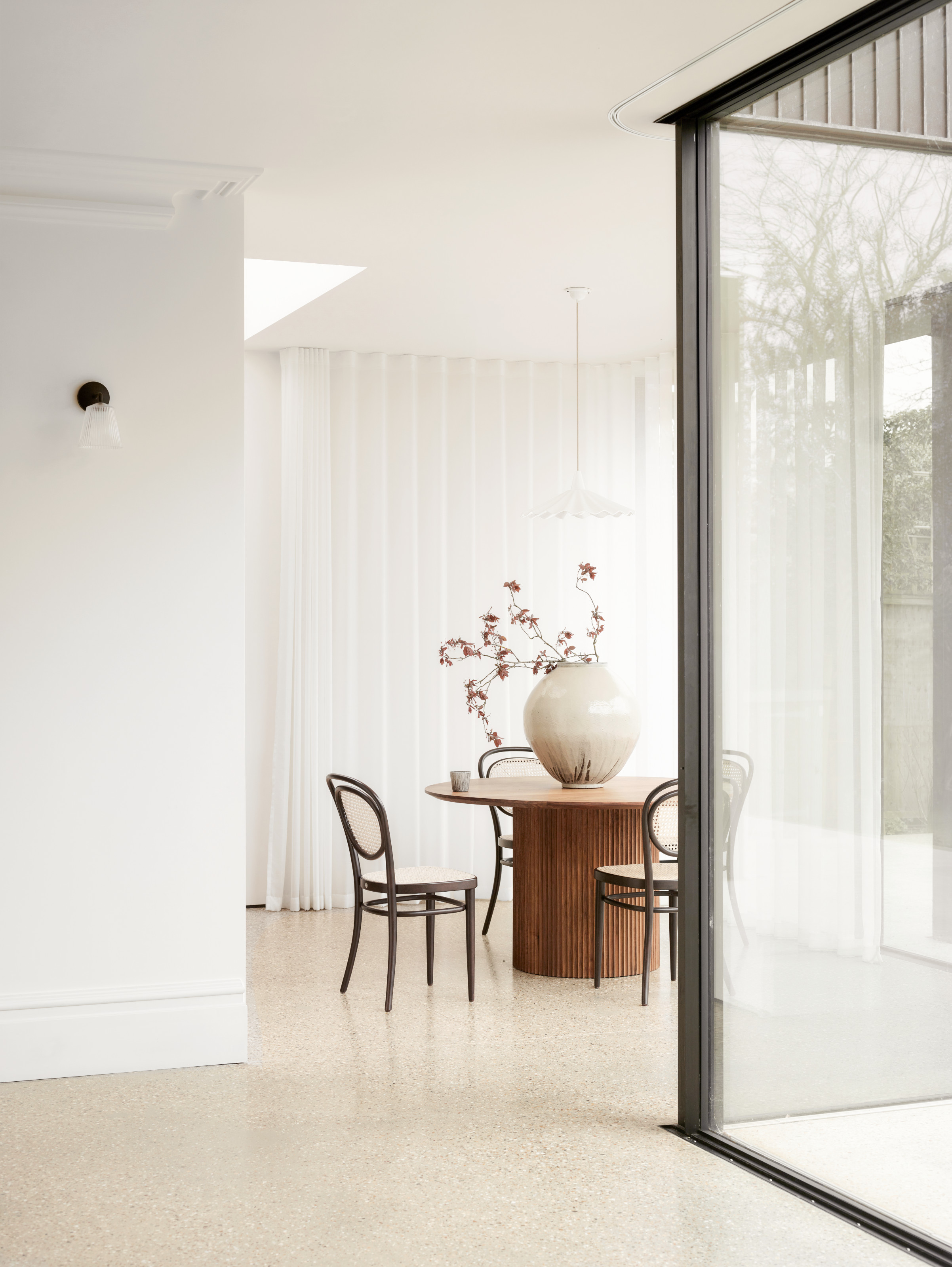 White dining area with sheer curtains, round wooden pedestal table and black and cane chairs