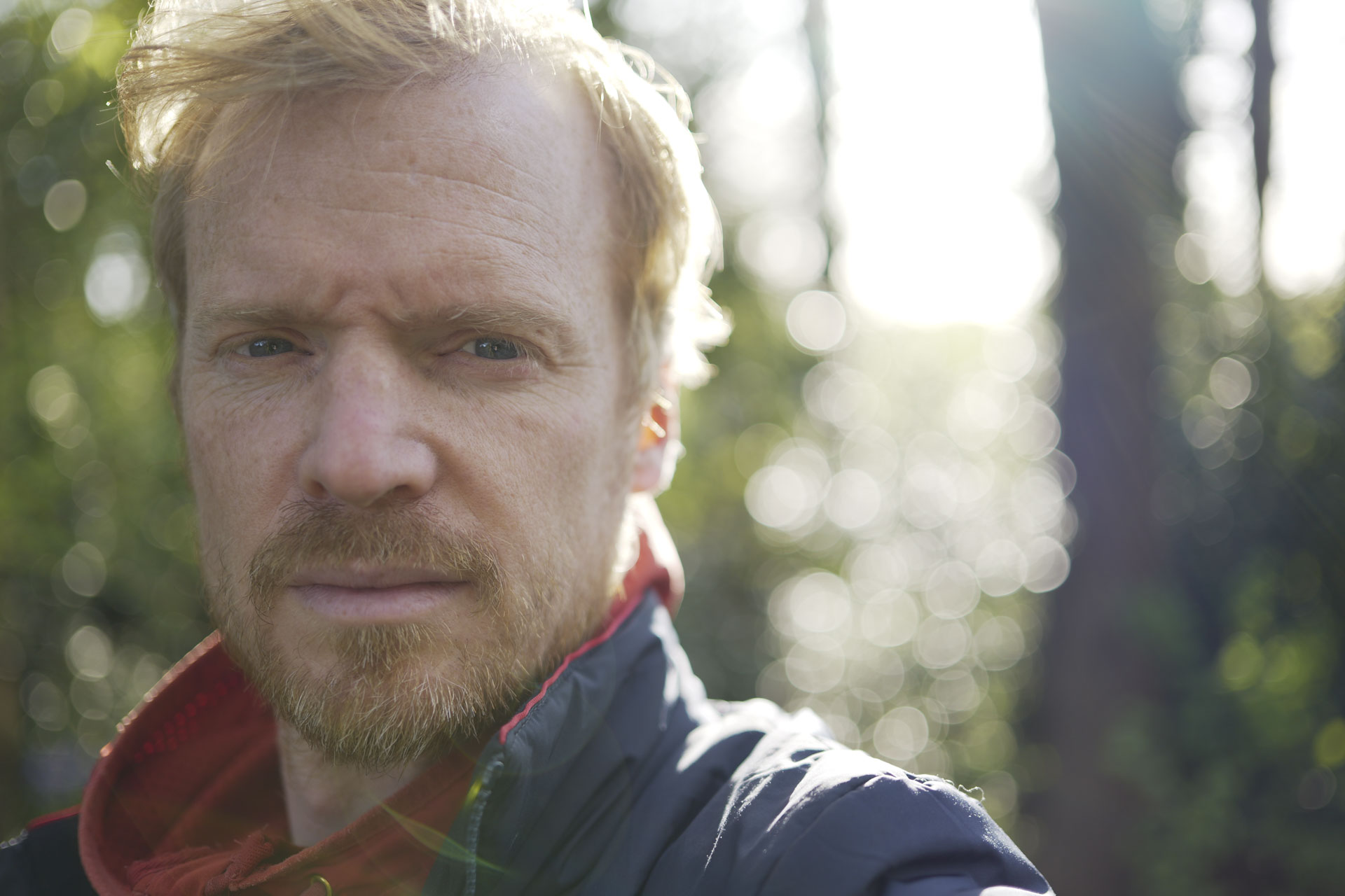 Man in an orange hoodie with strong sunlight behind him and dappled light through trees