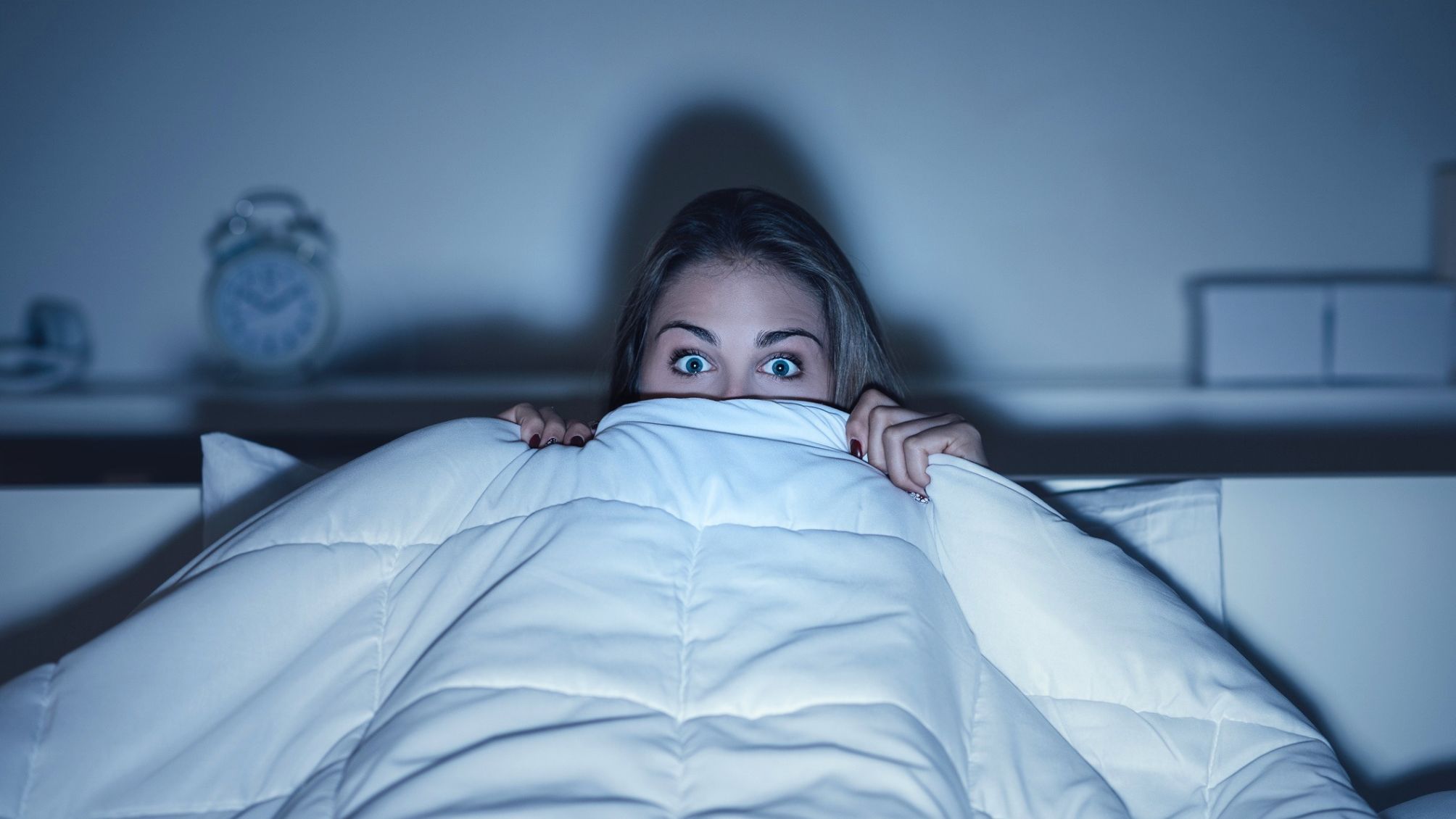 Woman watching a scary horror movie on tv late at night, she is frightened and hiding under the blanket