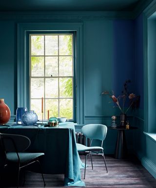 Striking teal painted dining room, walls and ceiling painted all one color, matching table cloth and upholstery, dark wooden flooring, black side table with flowers and decorative ornaments, candles and glass vases on dining table