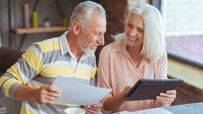 An older couple smile as they work on financial planning together with a tablet at their kitchen table.