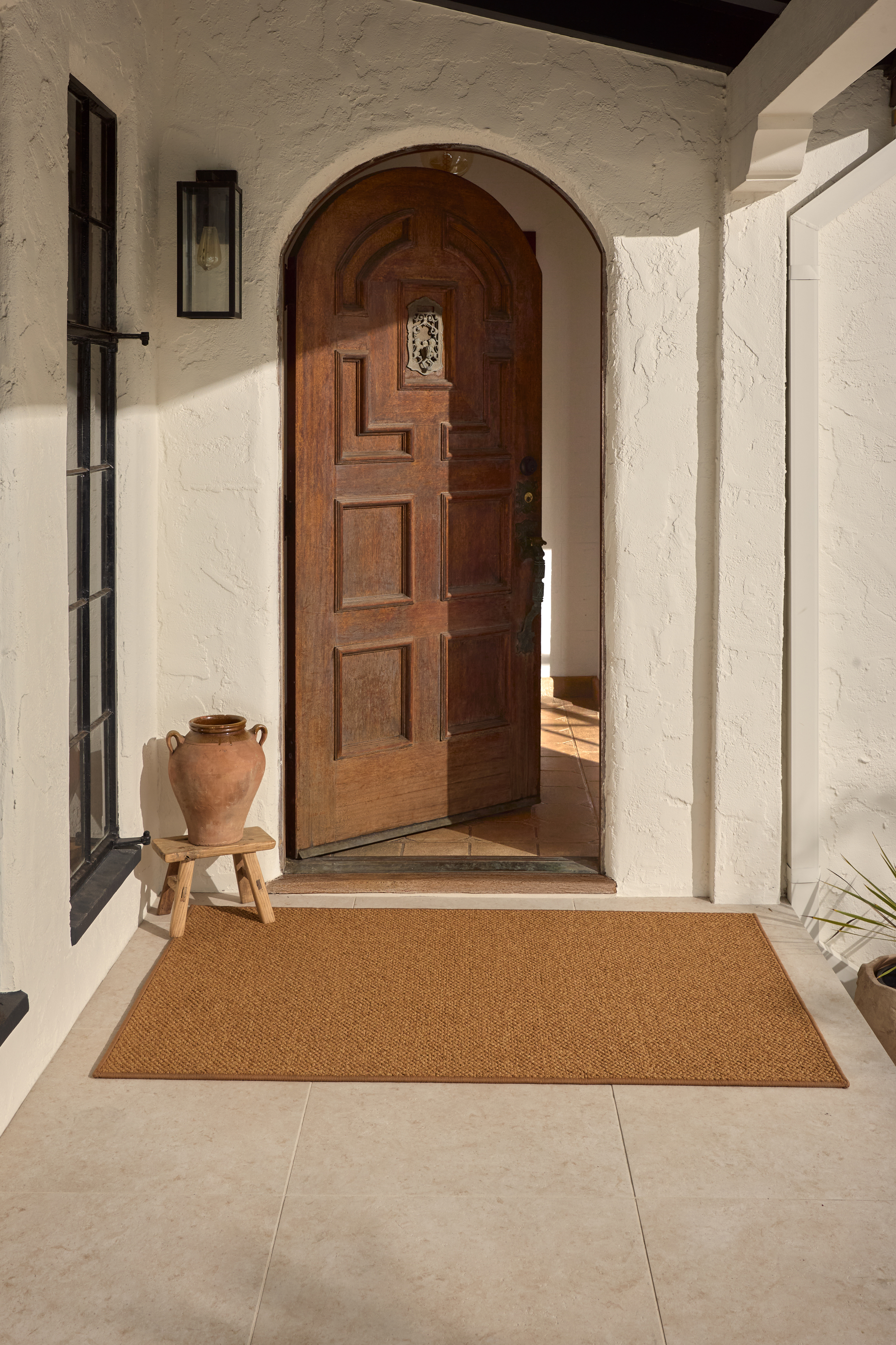 A dark jute-effect rug in an outdoor portico with white stucco walls and an old wooden door that is partly open. There is a wooden stool with a terracotta vase on it.