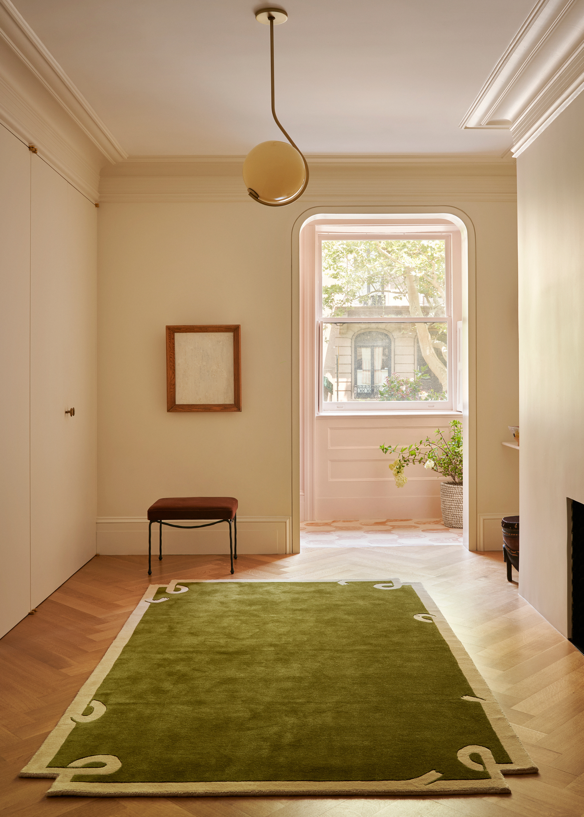 A bright room with a green rug, a brown stool, a hanging spherical pendant light, and a curved doorway leading to a large window