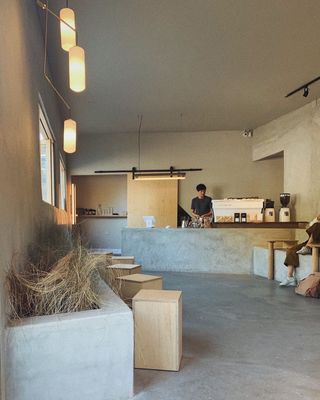 The brutalist, minimalist interiors of a coffee shop are molded our of bare stone, wood, and plants, with a coffee counter exclusively shaped out of a plank of stone.