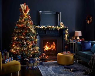A Christmas-themed living room with navy blue and copper decor including lit Christmas tree, mustard colored seat, traditional fireplace, blue patterned rug and blue velvet sofa