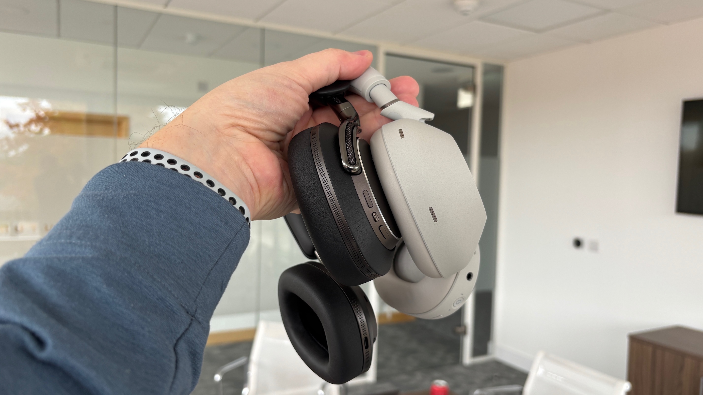 Bowers &amp;amp; Wilkins Px8 S2 and Sony WH-1000XM6 headphones held side by side in one hand in an office.