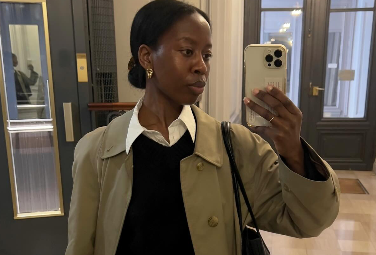 Woman wearing white button-down, black sweater, and car coat, taking a photo of herself in mirror.