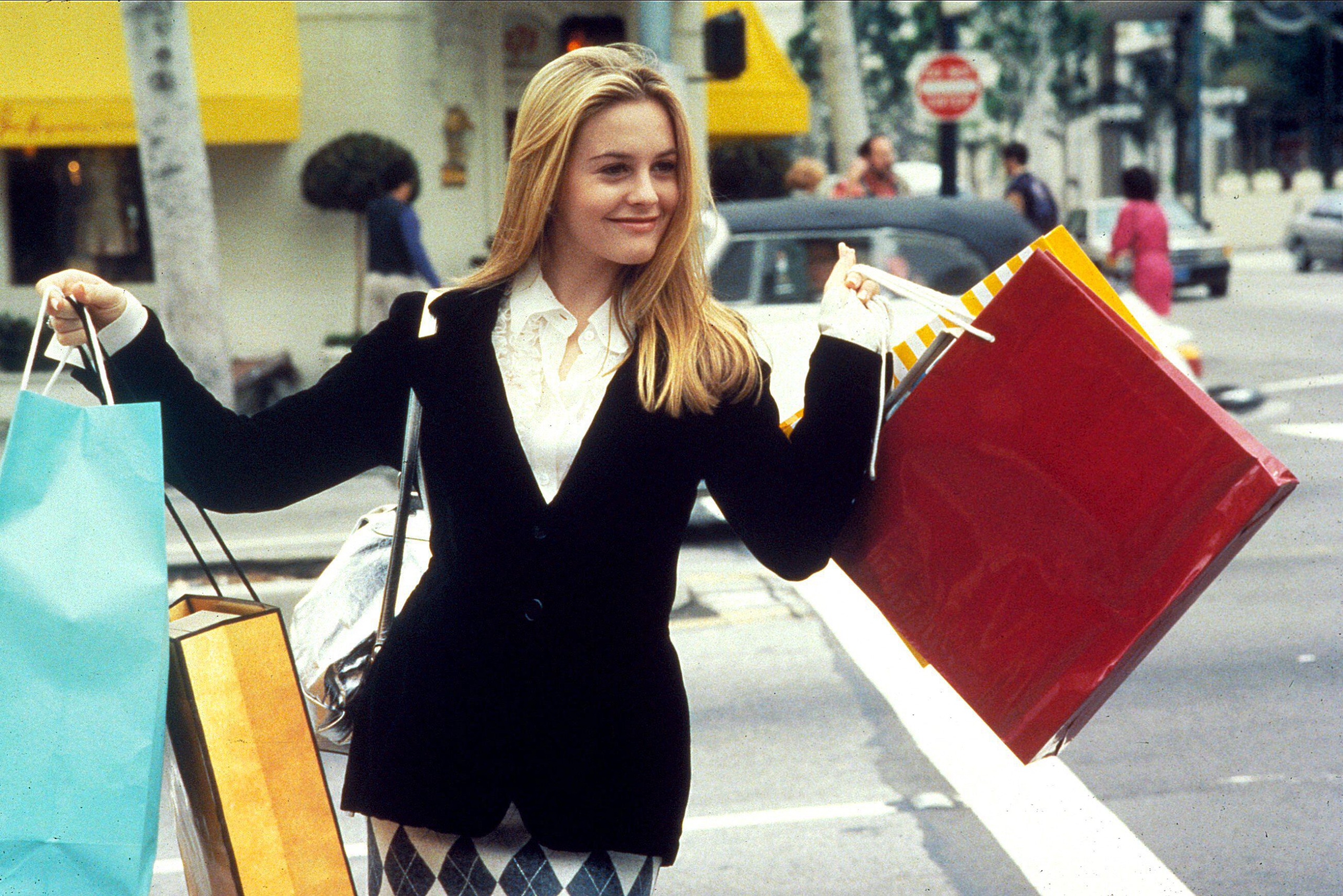 Alicia Silverstone as Cher in Clueless during the iconic shopping scene. She's wearing a white shirt and black blazer and clutching lots of colourful shopping bags. 