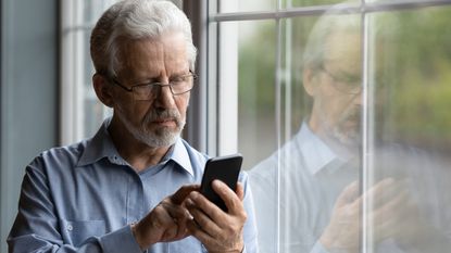 An older man looks concerned while looking at his smartphone next to a window.