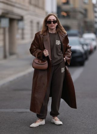 Sonia Lyson wearing Sporty & Rich brown cotton oversized logo sweater, matching Sporty & Rich joggers, a brown varnished leather long coat and Zara creamy white fluffy ballerinas