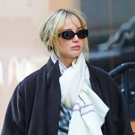 Jennifer Lawrence walking on the sidewalk in New York City wearing a black coat by The Row, a tied scarf, a plaid blue button-down shirt, lounge pants, yellow Polo socks, and New Balance 1906 sneakers.