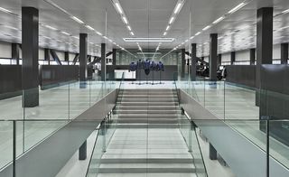 Interior view of the lobby at G-Star RAW headquarters, Amsterdam, white floor, centre stairwell, glass viewing barrier, dark grey pillars and beams, white tiled ceiling with strobe lighting, ground floor and upper floor desks and open plan office space, dark grey divider walls, tall glass windows, black floor standing body mannequins, with blue upper body clothing at the top of the stairs