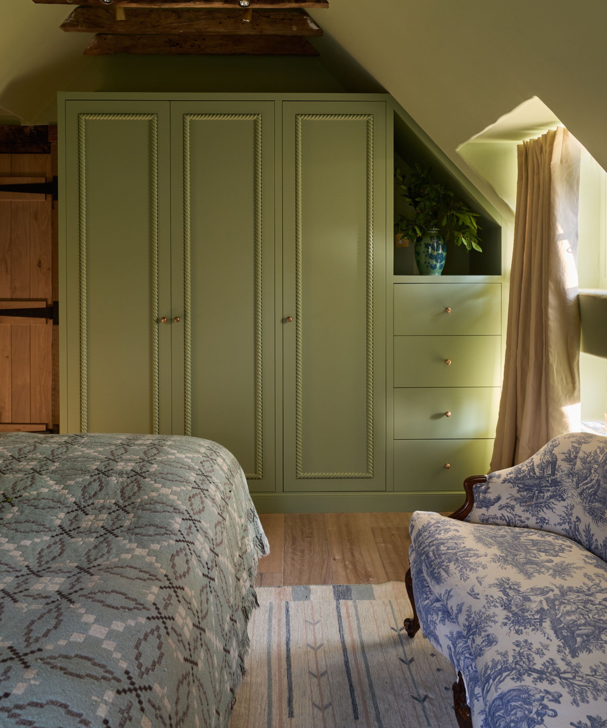 soft green cottage bedroom with sloped ceilings and bespoke cabinetry. a folk flatweave rug on the floor is paired with an antique settee