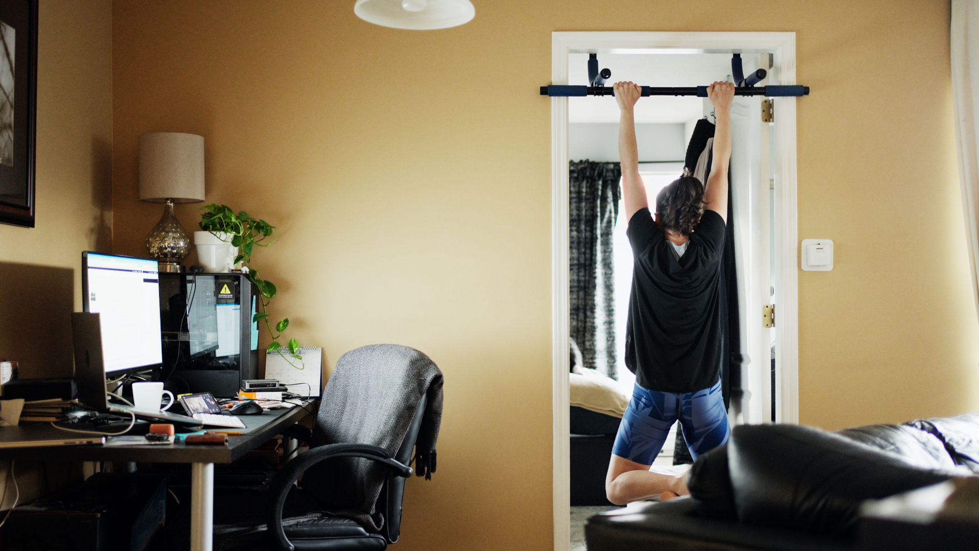 A person is seen doing pull-ups on a doorway bar in a home office space. The room is equipped with a desk and computer setup.