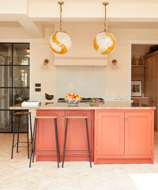A peachy pink kitchen island with a quartz surface with a fruit bowl on top, two white globe pendant lights above it, and three wooden stools tucked under it