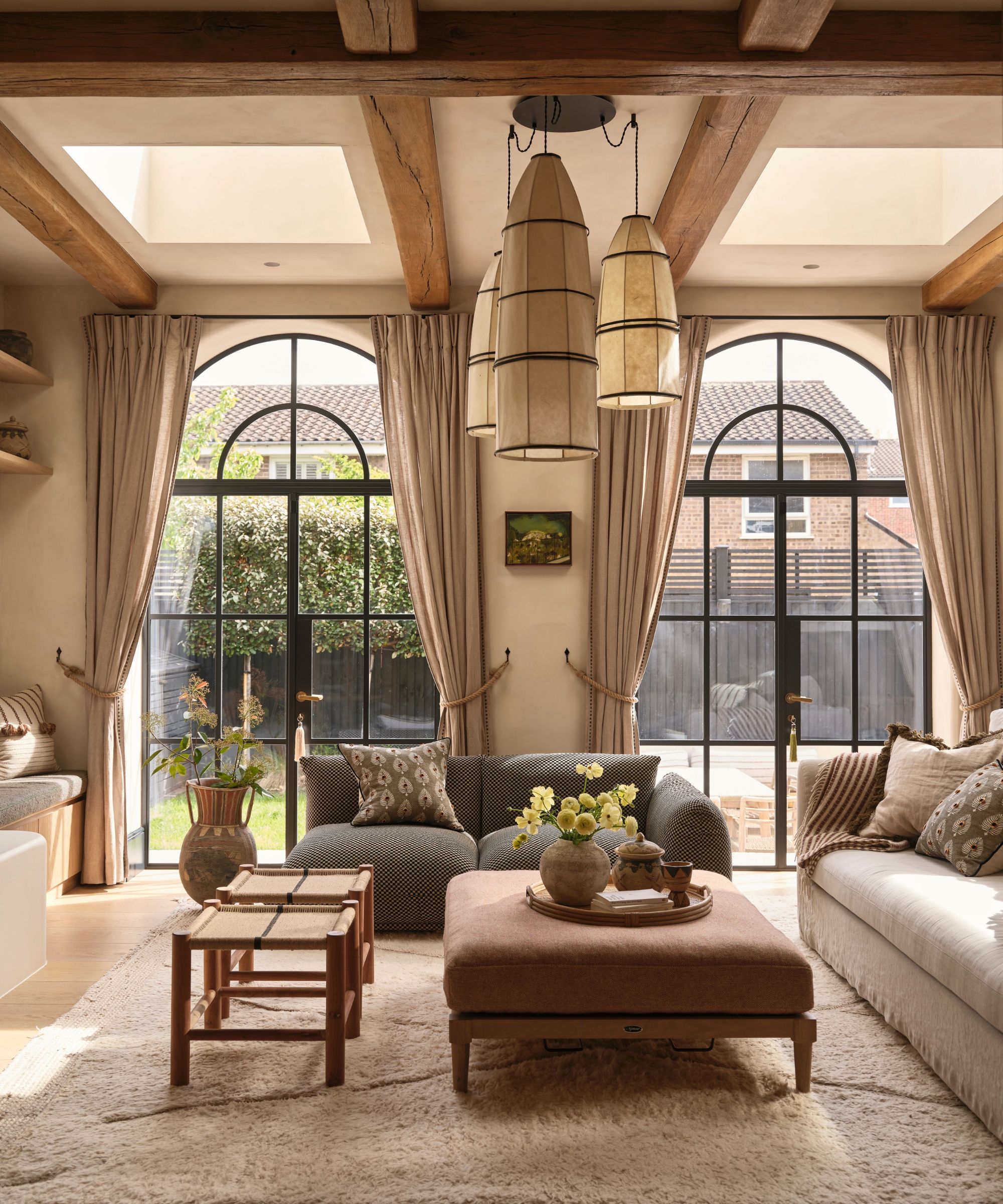 A bright living room with large windows and curtains, exposed wooden beams on the ceiling, and mismatched furniture with a grey couch, beige couch, burgundy ottoman, and large cream rug.