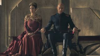 A regal woman in a red dress sat next to a bald man in a navy blue military dress-style outfit.