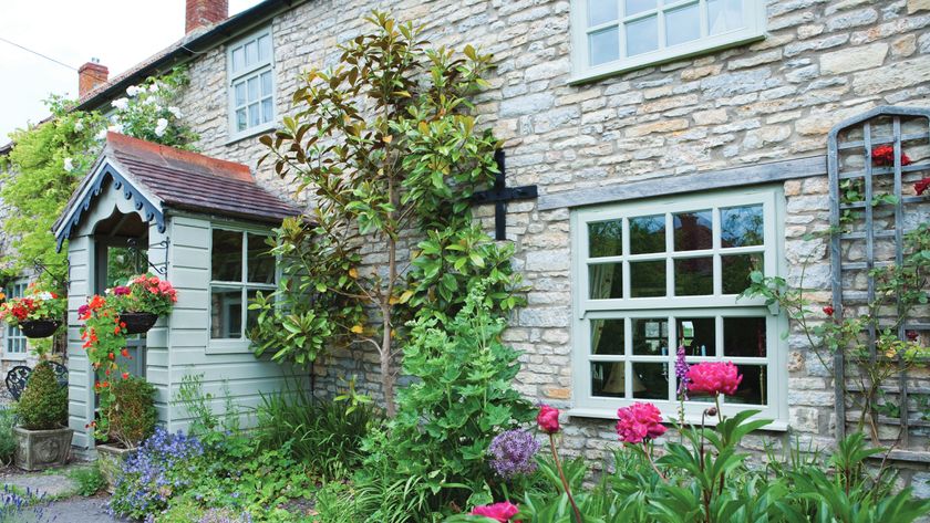 Exterior of stone house with green weatherboard clad porch, green windows, well planted border and trellis