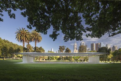MPavilion 2019 by Glenn Murcutt, surrounded by trees and grass, high skyscraper building stand in the distance with a blue sky