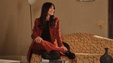 A woman in a terracotta orange suit with a black T-shirt and black sleek shoes sits atop a sculptural sofa with a wavy patterned texture in a beige-tinted room while staring to her left.
