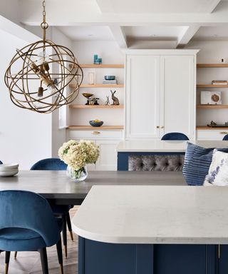A curved white kitchen island with a gray dining table in the middle with white flowers and blue velvet chairs eithe side, plus a brown woven pendant light above it