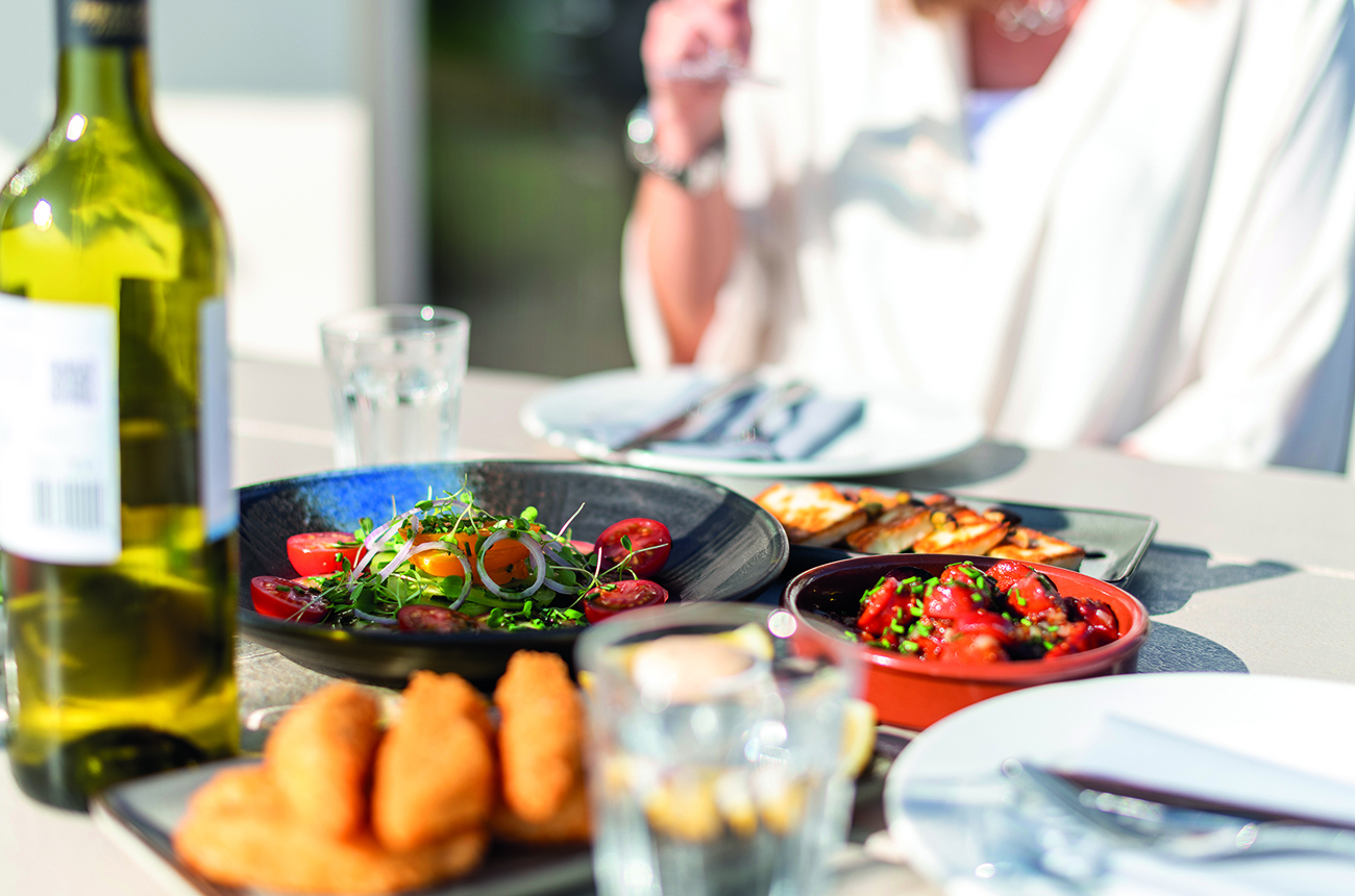 Summery plates of food including a colourful tomato salad laid out on a table outside with a bottle of white wine on a warm summers day.