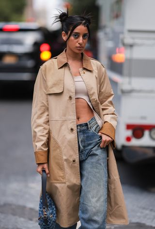 a street style photo of a woman wearing a vintage denim coach bag with jeans, a tan cropped tank top, and a trench coat