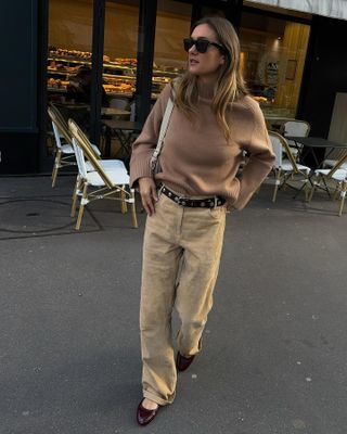 Anne wears a brown chunky sweater, brown stud belt, beige suede jeans, burgundy mary janes, and an ivory bag.