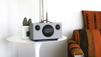 Audio Pro Addon T3+ in black and grey, sitting on white side table beside burnt orange sofa