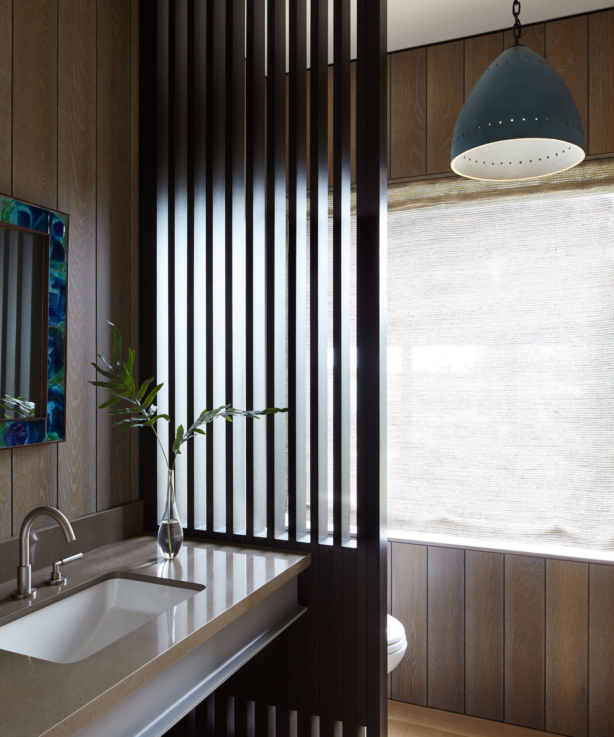 Contemporary bathroom with vertical wood slat partitions, a sleek tan vanity, and a blue pendant light against wood-paneled walls.