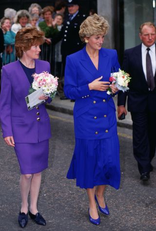 Princess Diana wears a royal blue skirt suit and carries a bouquet of flowers as she walks with her sister, Lady Sarah McCorquodale, who is wearing a purple skirt suit