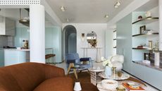 a modern living room with 70s style, mint blue cabinetry, a rust orange sofa, blue velvet armchair, marble nesting coffee tables, shaggy armchair, display shelves, and a perforated surface along the roofline concealing aircon