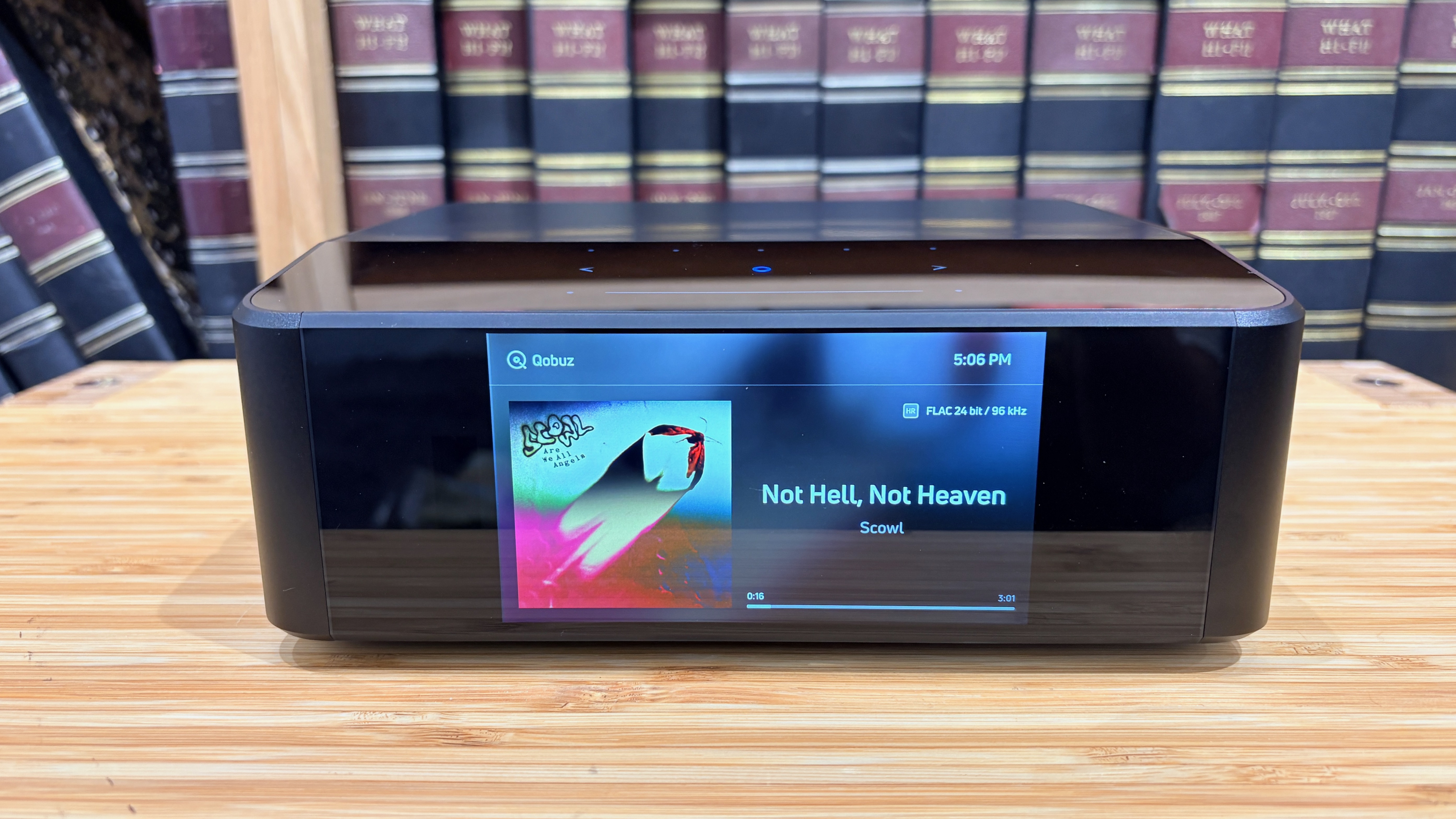 Bluesound Node Icon music streamer on wooden hi-fi rack in front of bookcase, with album artwork showing on screen