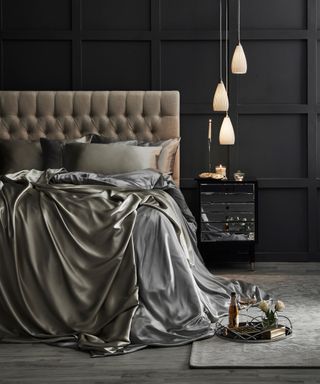 A black bedroom with wall paneling and silk bedding, trio of pendant ceiling lights and mirrored sideboard