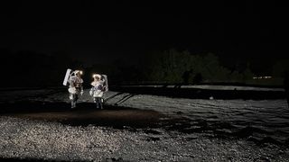 two people stand on a gravel landscape wearing spacesuits.